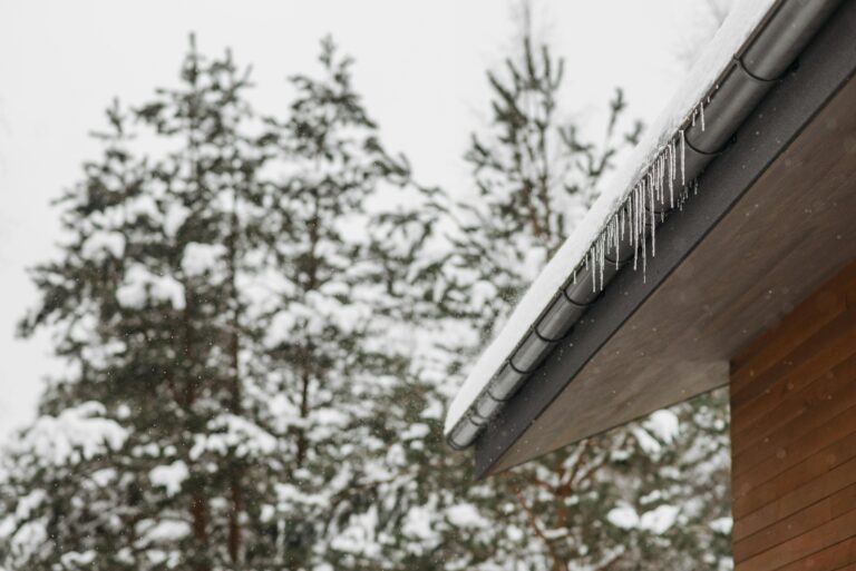 Stunning winter scene of icicles hanging from a snow-covered roof with snowy pines in the background.